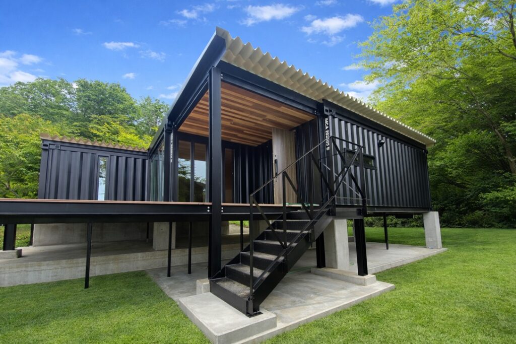 Modern black shipping-container home on stilts with a metal staircase and wooden deck, on a green lawn.