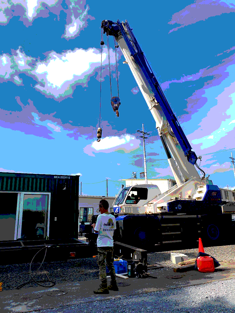 Large white-and-blue mobile crane lifts a hook and cables at a worksite while a worker in a Patagonia shirt watches nearby.